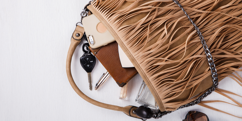 Handtasche mit Fransen, Smartphone, Autoschlüssel, Parfumflakon, Kugelschreiber, Damenaccessoires auf hellem Untergrund