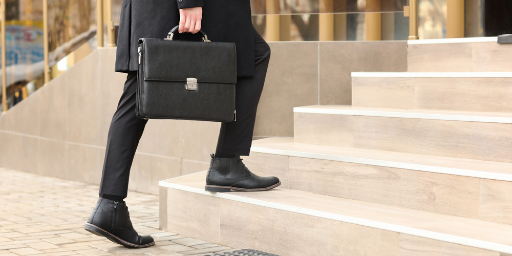 Mann in schwarzer Hose und Stiefeletten, hält schwarzen Aktenkoffer, steigt Treppe hoch