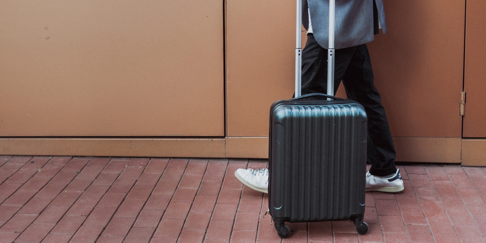 schwarzer kleiner Koffer, helle Sneakers, orangene Wand, kompakt, unterwegs