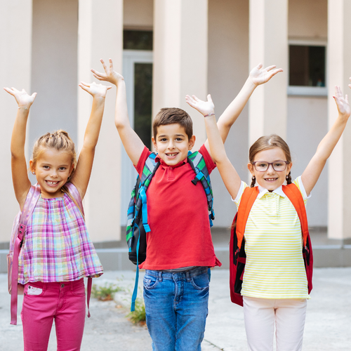Schule, fünf Kinder mit bunten Schulrucksäcken, halten die Hände in die Luft, stehen vor einem Gebäude, fröhliche Stimmung, Schulbeginn