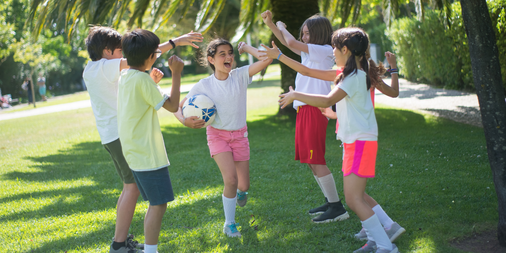 Sechs Kinder im Park, ein Mädchen mit Fußball in der Hand, fünf Kinder stehen jubelnd um das Mädchen, Sportbekleidung, sonnig, ausgelassene Stimmung