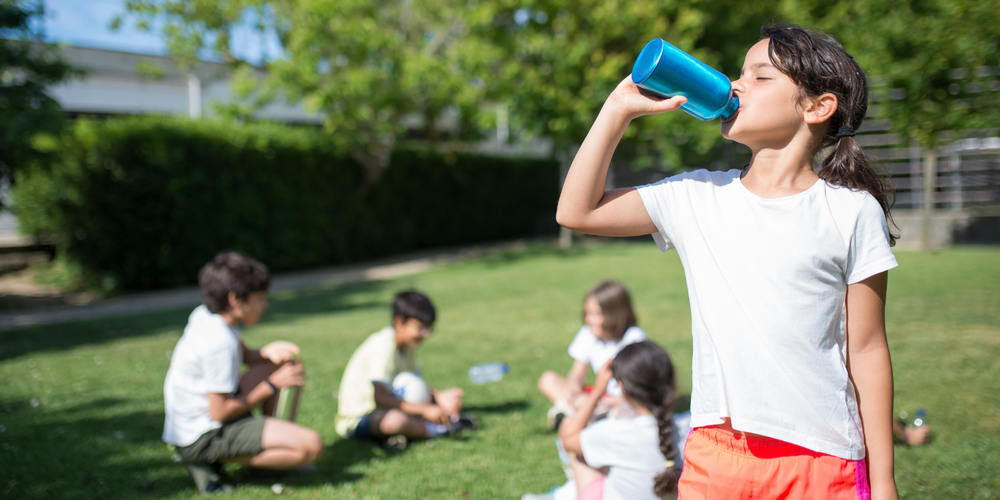 Trinkflaschen, Mädchen in weißem Shirt trinkt aus blauer Flasche, Wiese im Hintergrund, andere Kinder sitzen auf dem Rasen