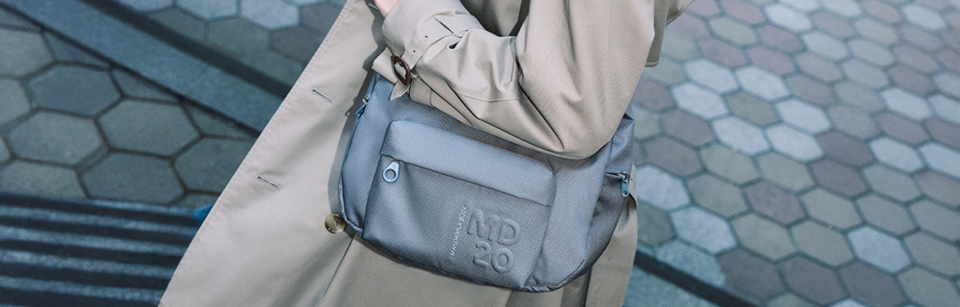 Mandarina Duck, Umhängetasche, grau, sportlich-modern, getragen über der Schulter, Trenchcoat, urbaner Hintergrund, Pflastersteine
